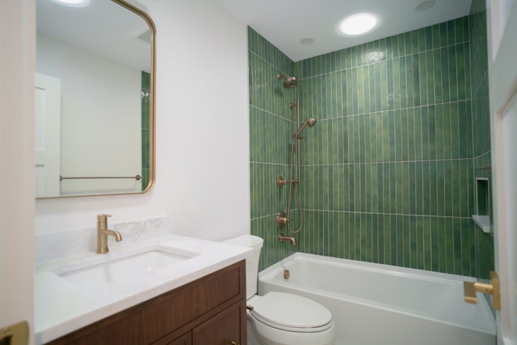 Remodeled bathroom with green vertical tile shower, brushed brass fixtures, a toilet and a walnut vanity